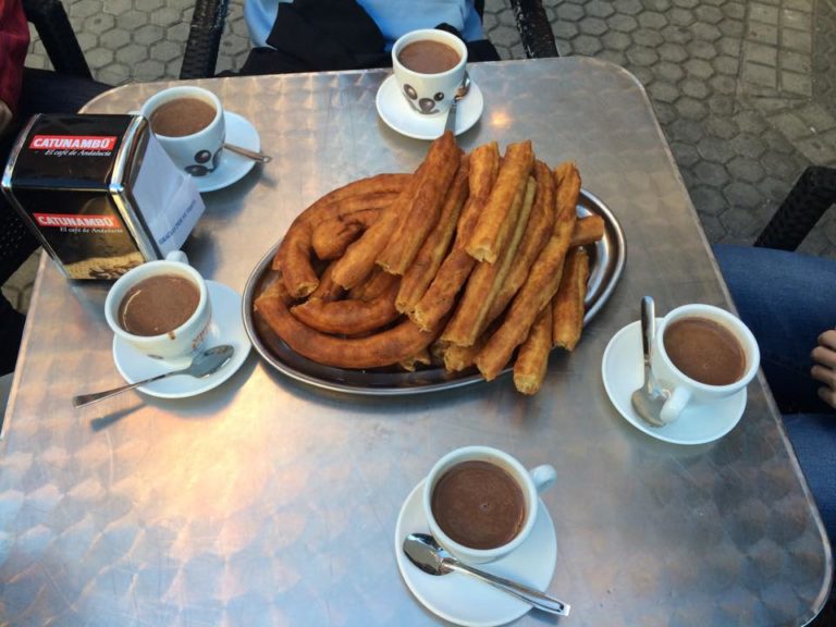 Pile of churros on a plate with five mugs of chocolate on a table