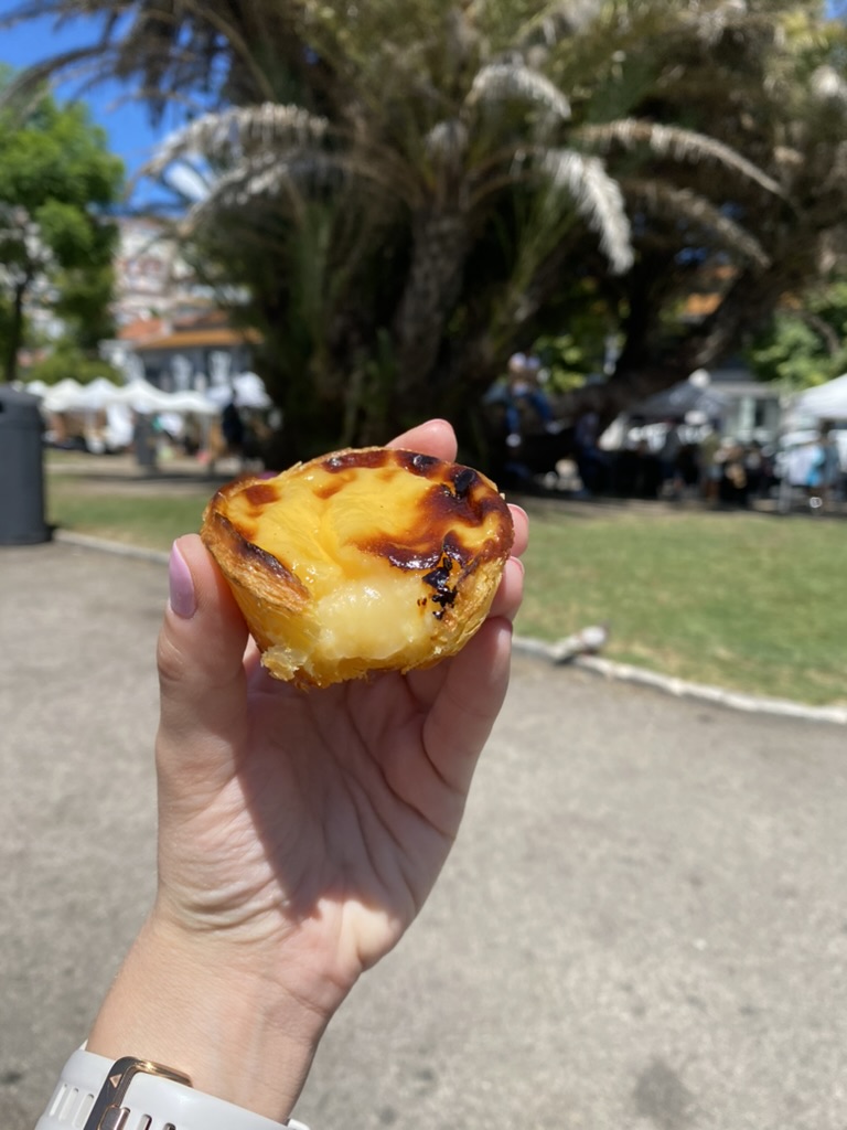 Hand holding pastel de nata with a bite taken out of it with a park in the background