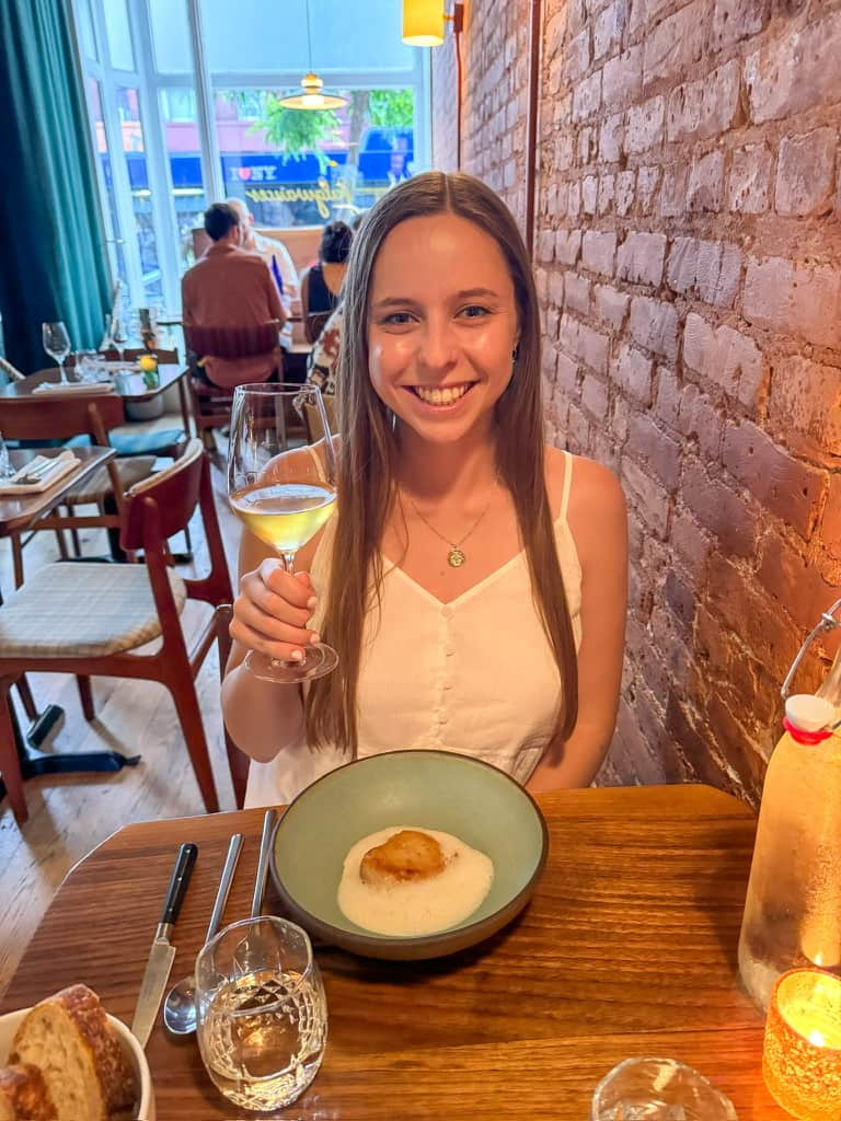 Woman smiling holding up a glass of white wine with a small plate of food in front of her