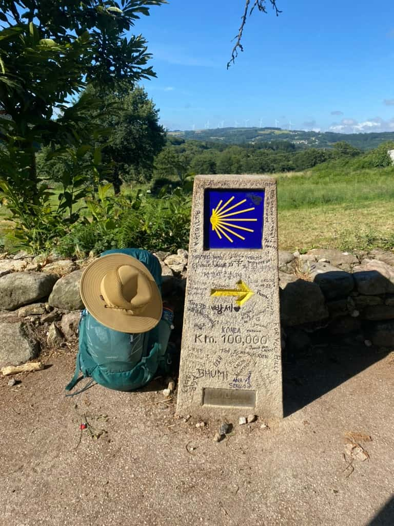 Camino Francés 100KM remaining marker with a hiking pack and mountains in the background