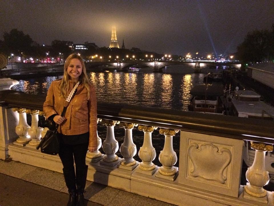 Woman smiling at night on a bridge in Paris, France with the Eiffel Tower in the background.