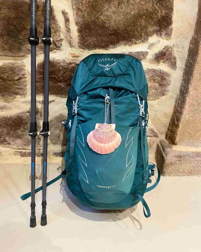 Green Osprey hiking pack with a shell hanging from it and hiking poles next to it leaning against a stone wall.