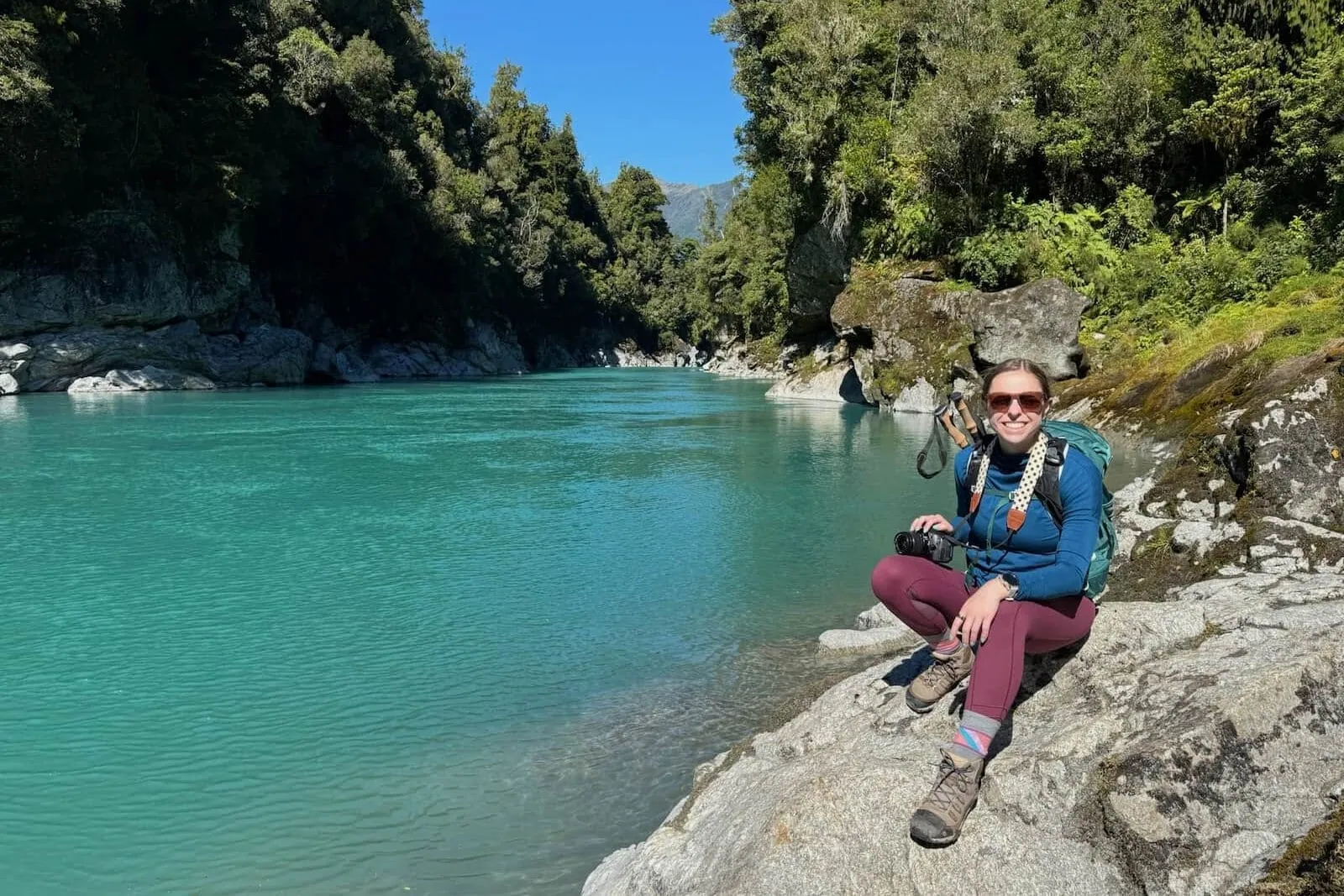 Mikala sitting smiling on a rock in front of Hokitika Gorge in New Zealand