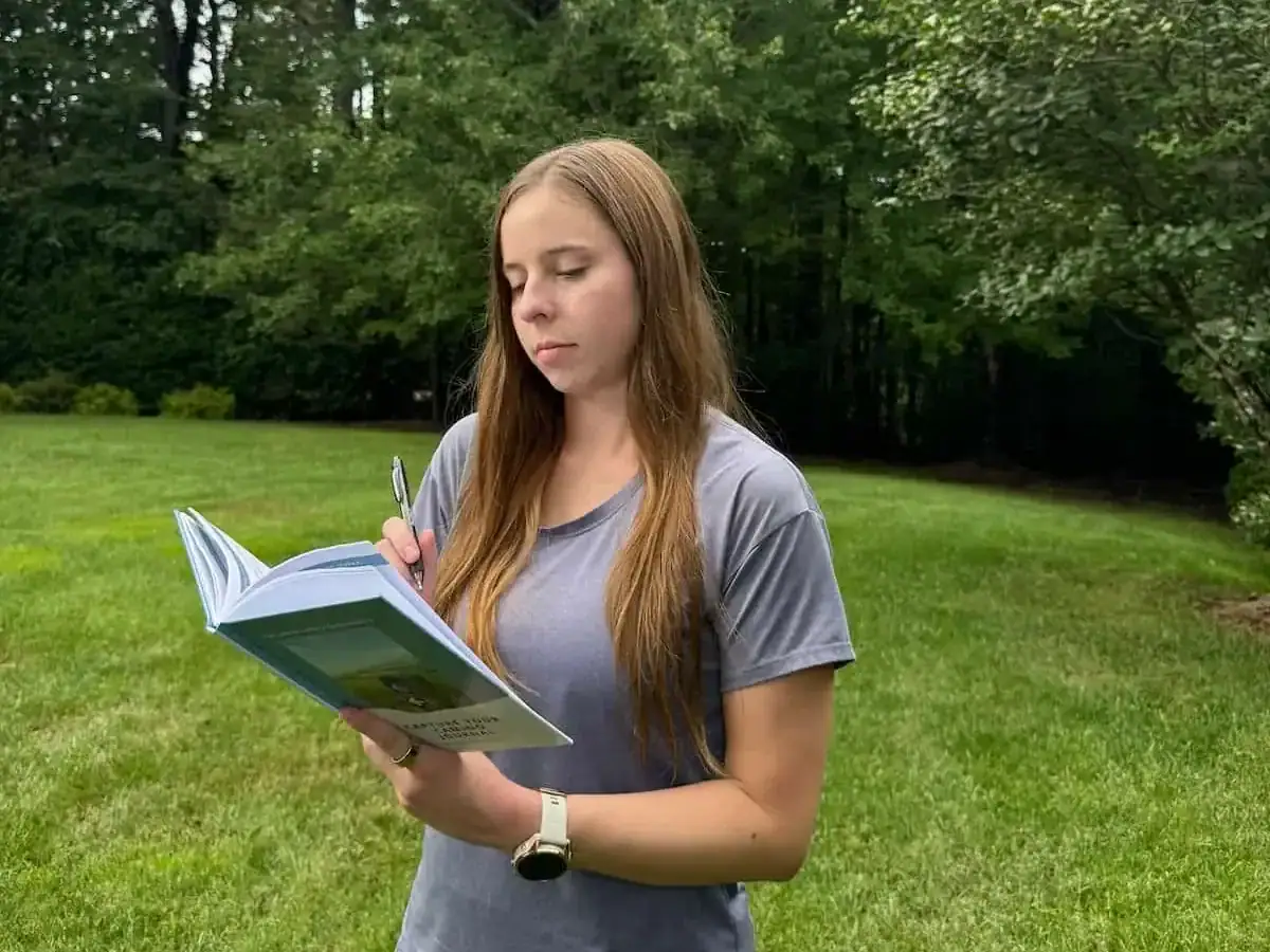 Mikala looking down at a small journal with green grass and a forest behind her